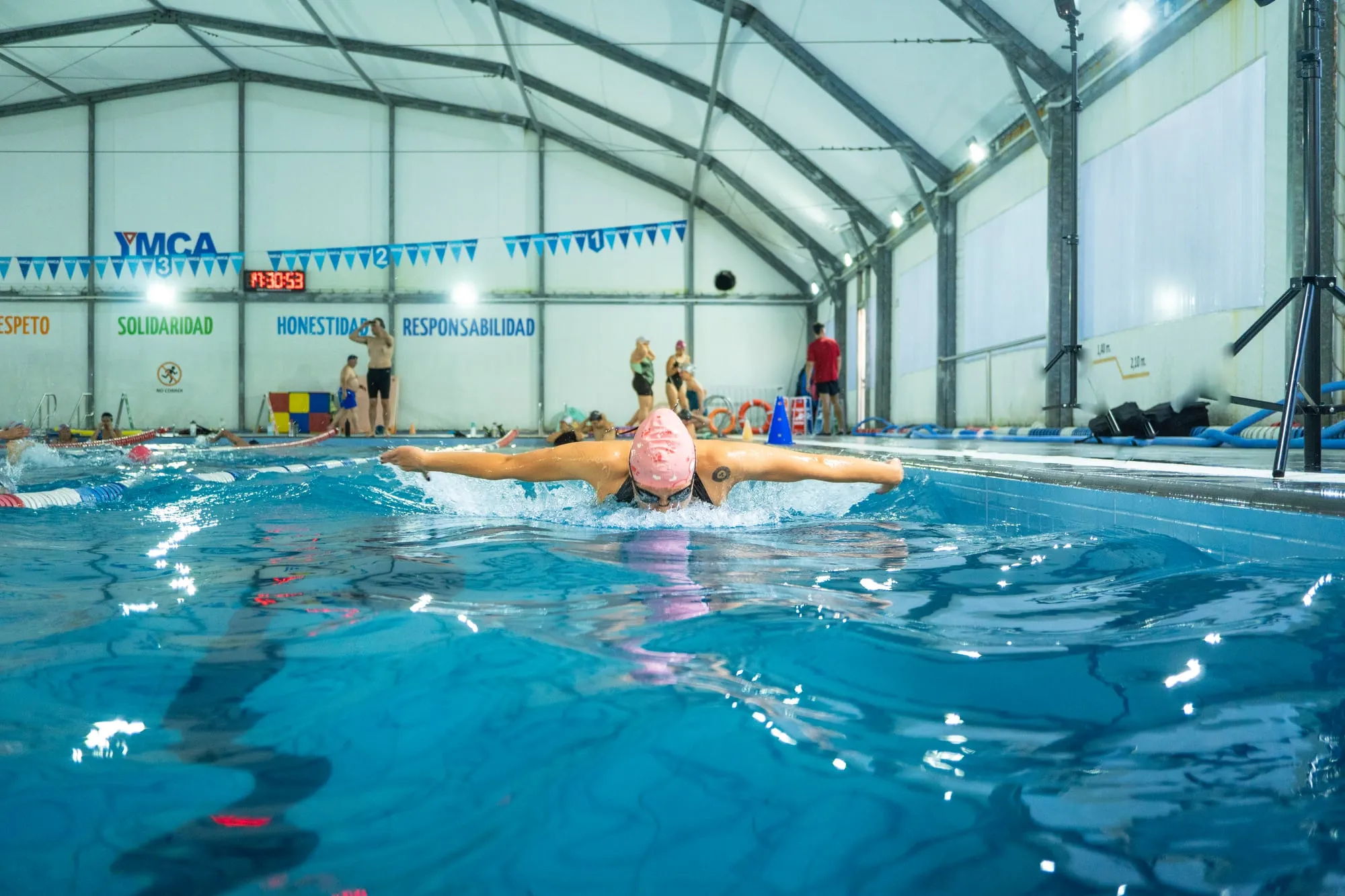 Nadadora con gorra rosada avanza en estilo mariposa por una piscina techada, bajo banderines y letreros de valores