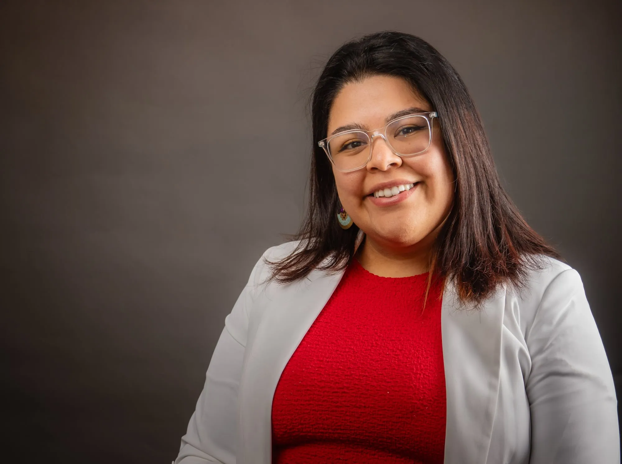Mujer sonriente con lentes, blusa roja y chaqueta blanca en un retrato de estudio sobre fondo oscuro
