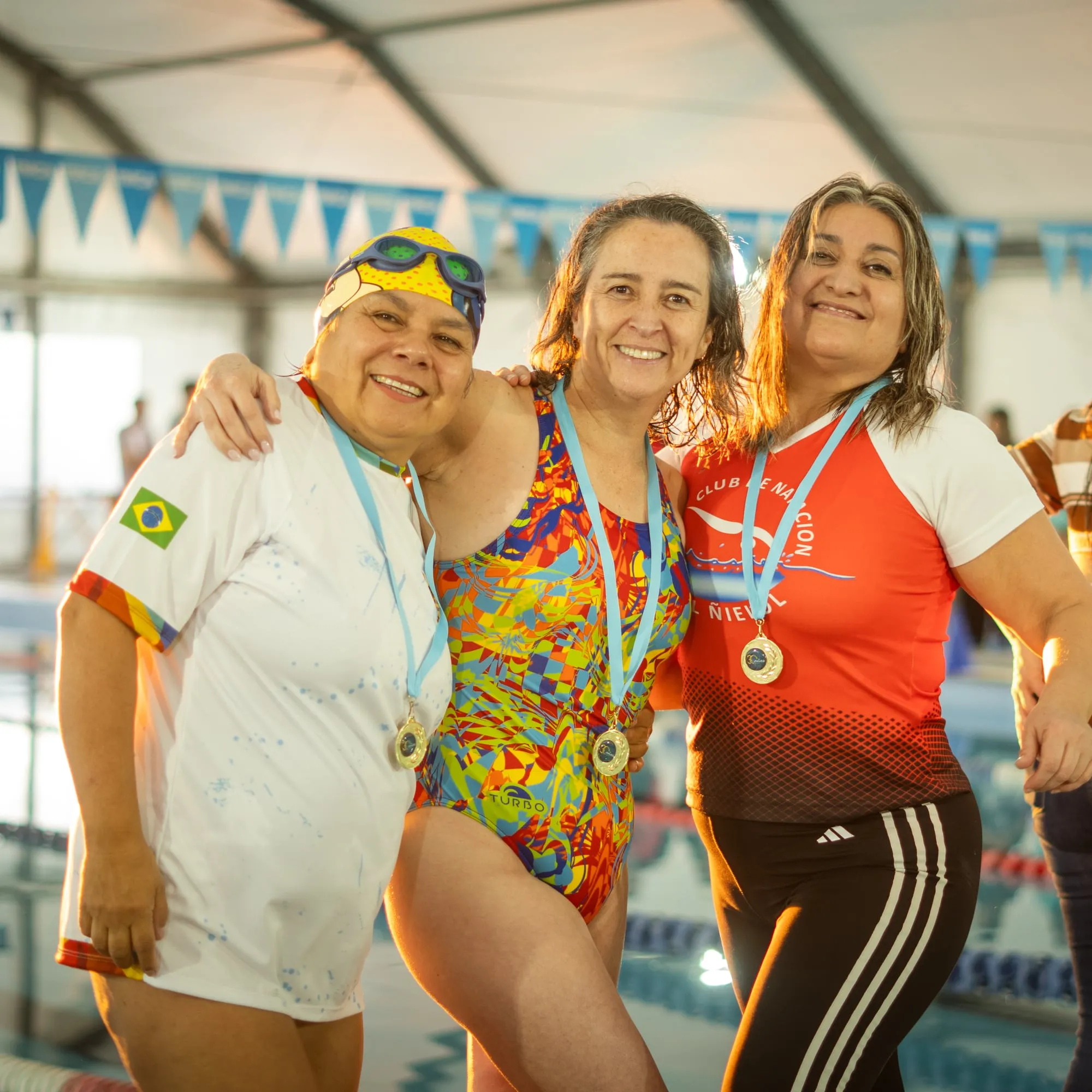 Tres nadadoras abrazadas y sonrientes muestran sus medallas al borde de la piscina