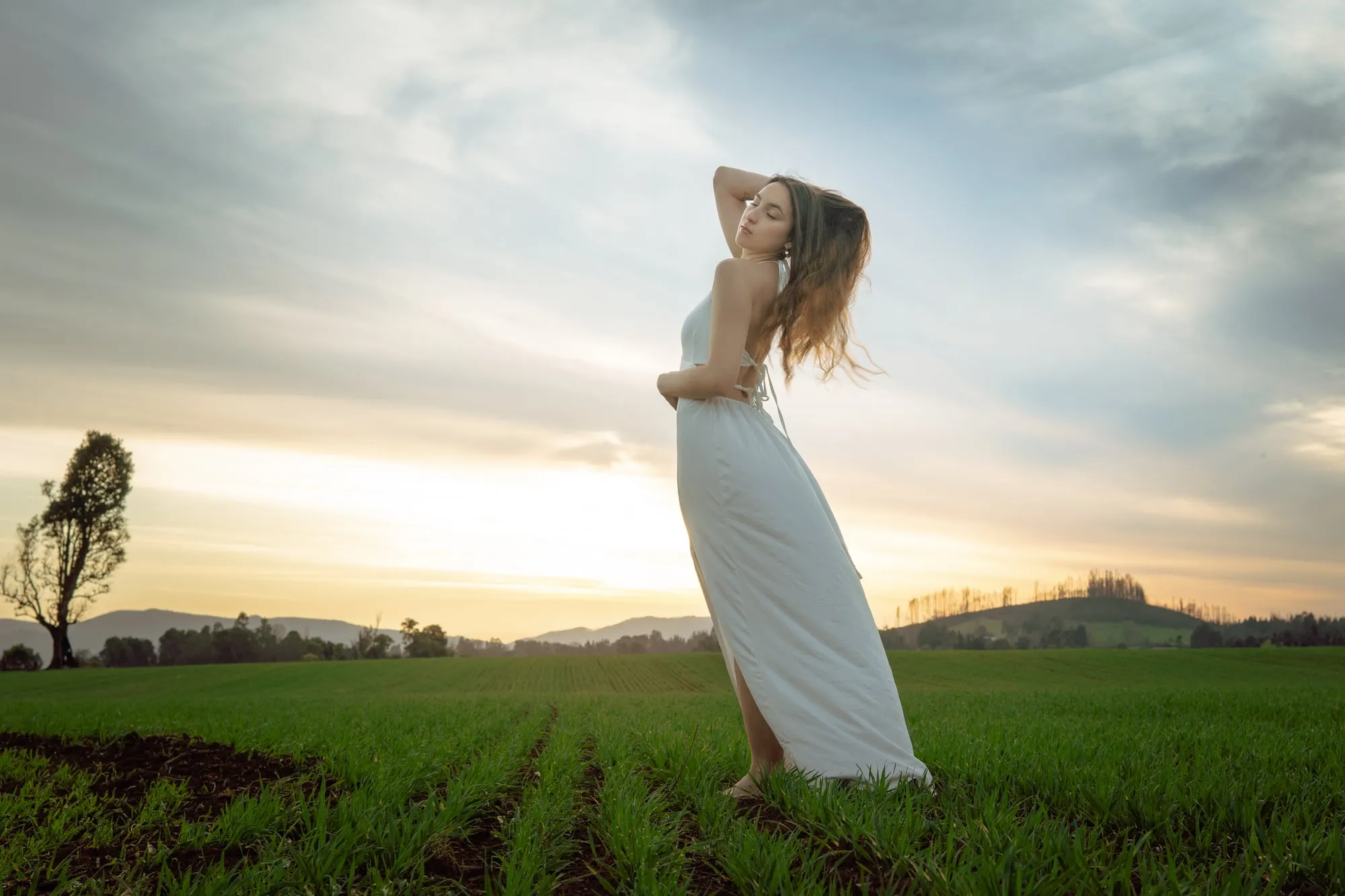 Mujer con vestido blanco de pie en un campo sembrado, con el cielo del atardecer y cerros al fondo