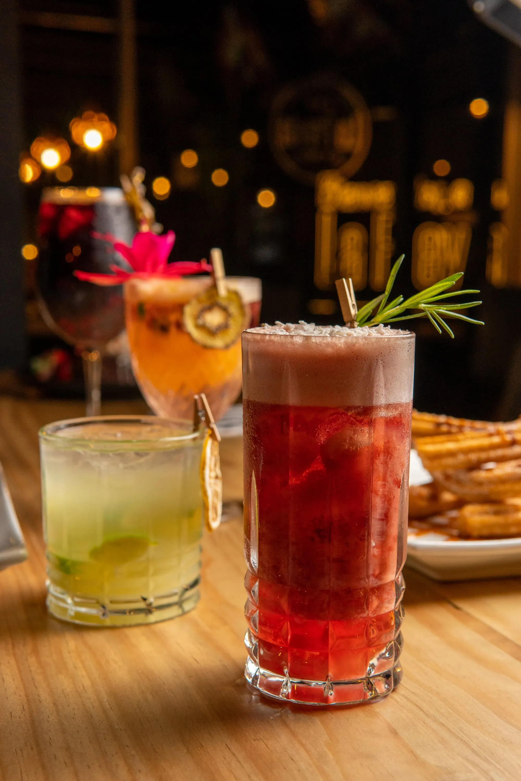 Cócteles de colores con fruta y romero sobre una mesa de madera, con las luces del bar desenfocadas detrás