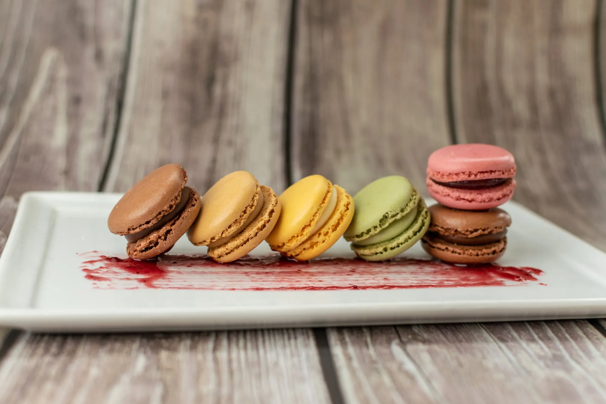Macarons de cinco colores en fila sobre un plato blanco con un trazo de salsa de frutos rojos